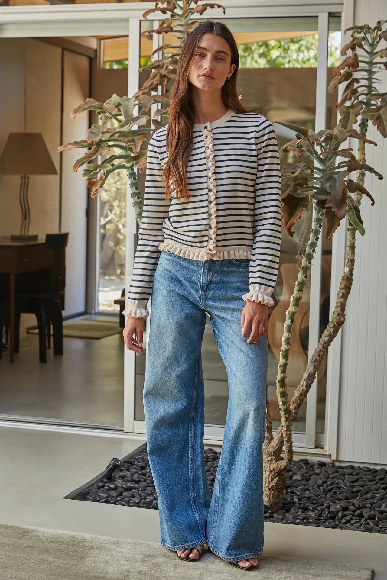 A woman stands in a modern home entryway wearing a cream and navy blue horizontally striped knit cardigan with ruffled trim, paired with wide-leg blue jeans.