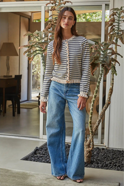 A woman stands in a modern home entryway wearing a cream and navy blue horizontally striped knit cardigan with ruffled trim, paired with wide-leg blue jeans.