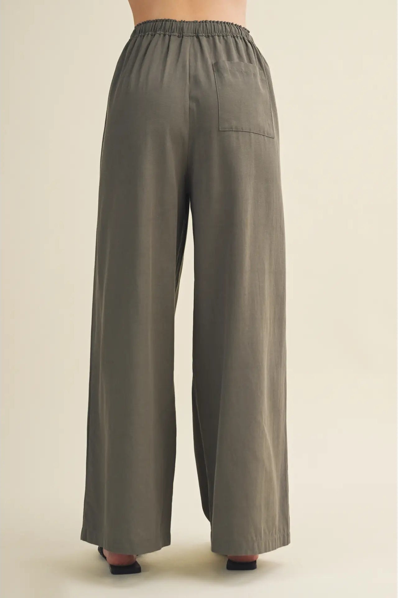 A person shown from the back wearing wide-leg trousers in a muted olive-gray color, photographed against a plain light beige studio background.