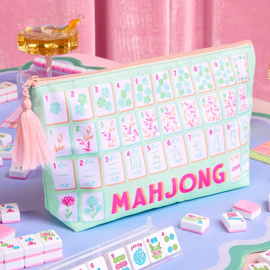 A mint green zippered pouch designed to look like a mahjong board, printed with various mahjong tiles and the word 'MAHJONG' in pink. The pouch has a pink tassel zipper pull and is shown on a styled tabletop with other miniature mahjong tiles.