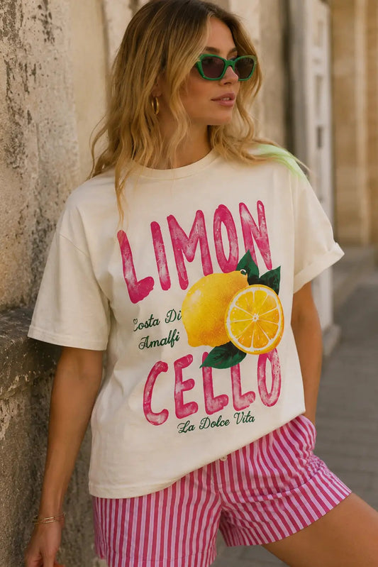 Woman wearing a t-shirt with 'Limon Cello' design and pink shorts, standing against a stone wall.