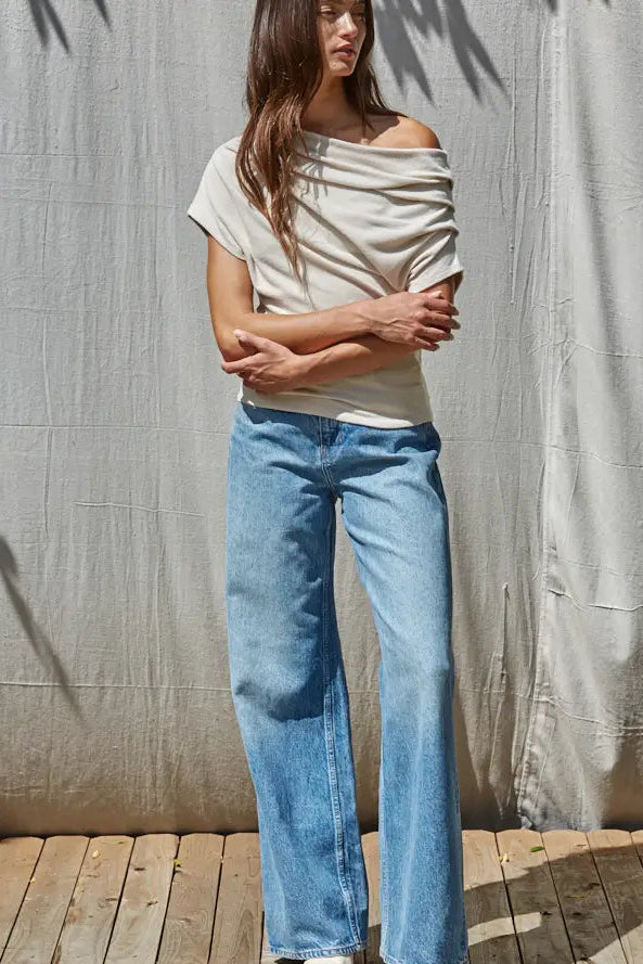 A woman wearing a light beige off-the-shoulder short-sleeve top and light blue wide-leg jeans, standing against a textured beige fabric backdrop on a wooden deck.