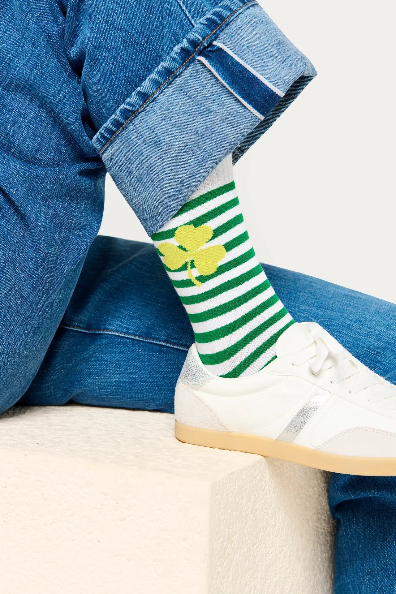 Person wearing green and white striped socks with a yellow clover design, paired with blue jeans and white sneakers.