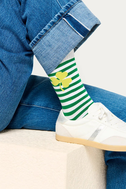 Person wearing green and white striped socks with a yellow clover design, paired with blue jeans and white sneakers.