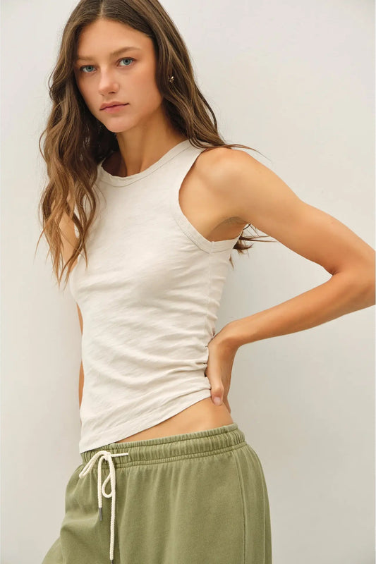 A woman modeling a sleeveless crew-neck tank top in off-white, paired with olive green drawstring sweatpants, against a plain light gray background.