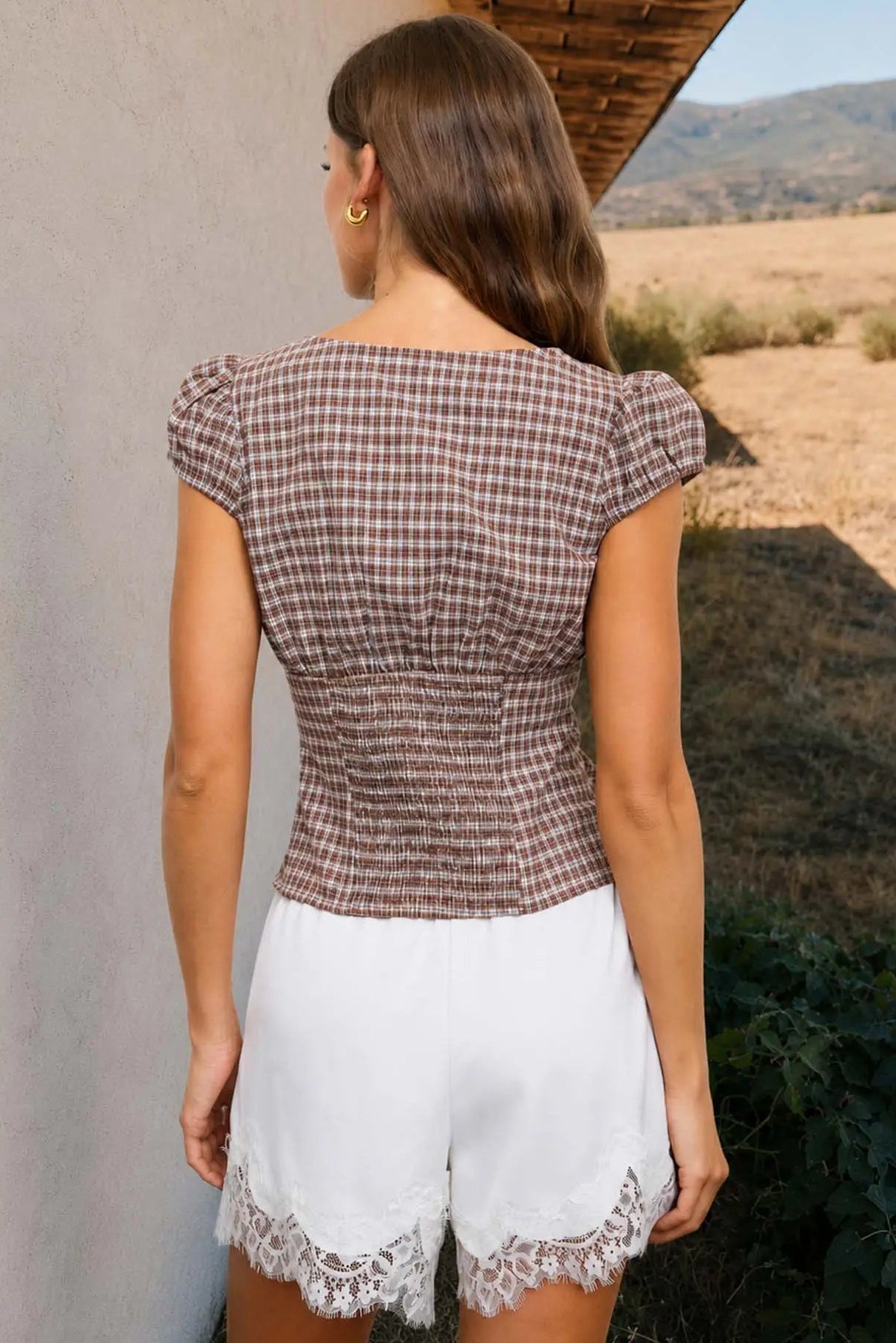 A woman shown from the back wearing a brown and white checkered puff-sleeve top and white shorts with a lace hem, standing outdoors next to a wall.
