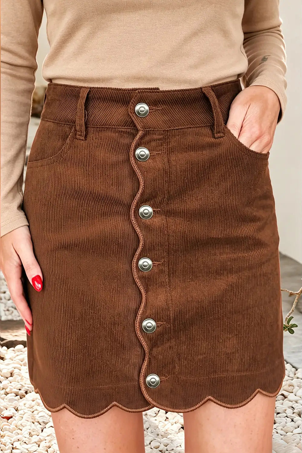 A brown corduroy mini skirt with a scalloped button-front hem, worn by a model in a beige long-sleeve top. The skirt features silver-tone buttons and side pockets.