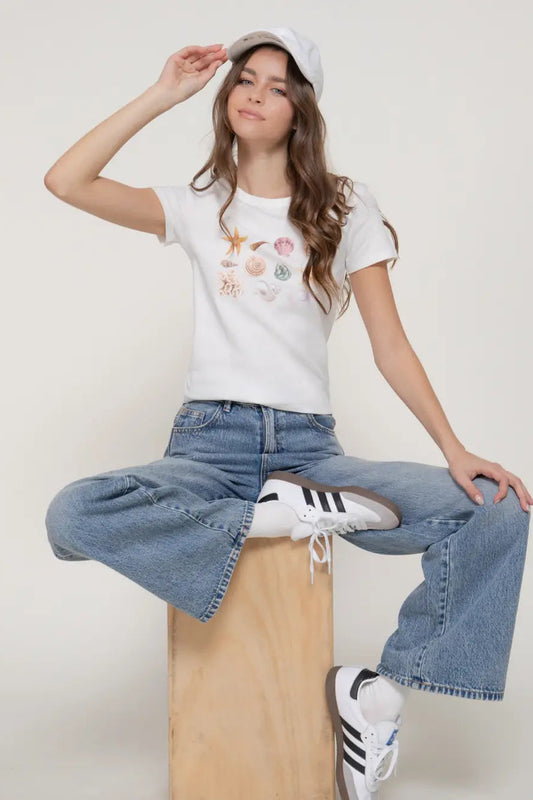 Person wearing a white t-shirt with floral designs, blue jeans, and white sneakers on a light background