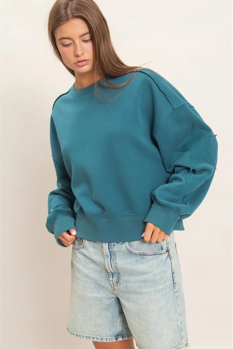 A woman wearing a teal blue oversized crewneck sweatshirt and light-wash denim shorts, standing against a plain beige studio background.