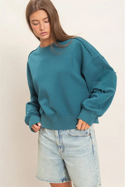 A woman wearing a teal blue oversized crewneck sweatshirt and light-wash denim shorts, standing against a plain beige studio background.