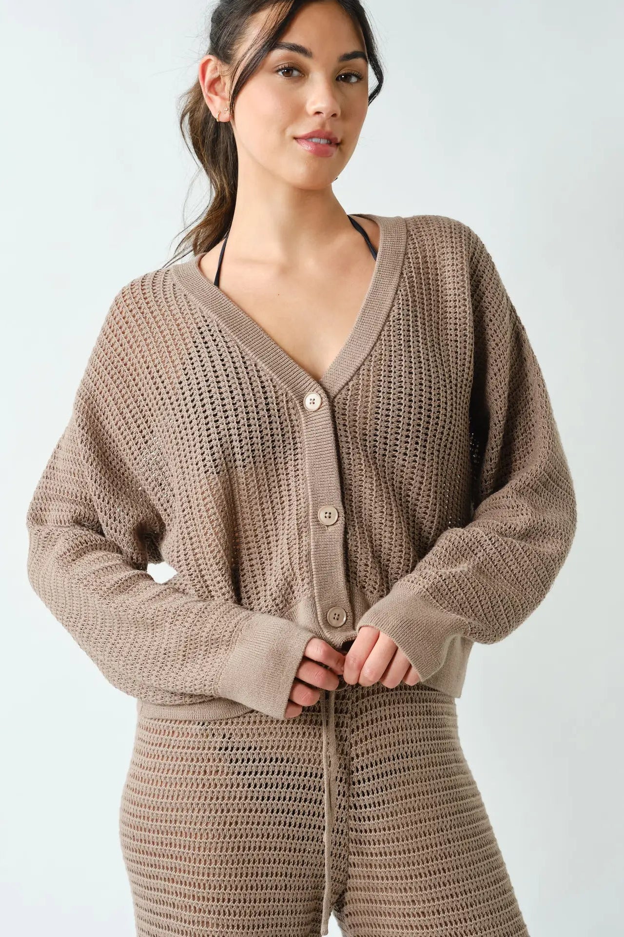 A woman wearing a chunky knit cardigan and matching knit pants in a taupe brown color, photographed against a light gray studio background.