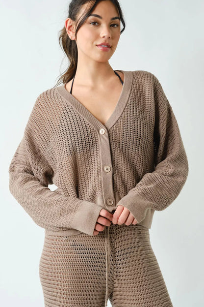 A woman wearing a chunky knit cardigan and matching knit pants in a taupe brown color, photographed against a light gray studio background.
