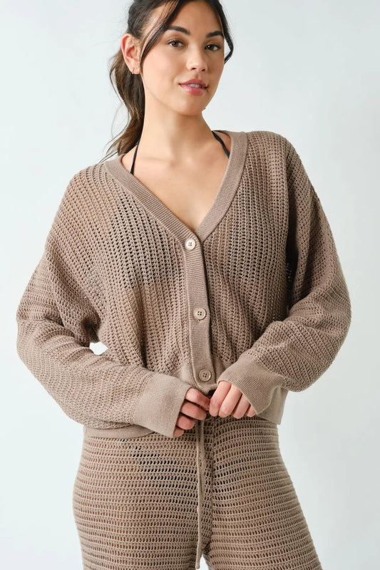 A woman wearing a chunky knit cardigan and matching knit pants in a taupe brown color, photographed against a light gray studio background.