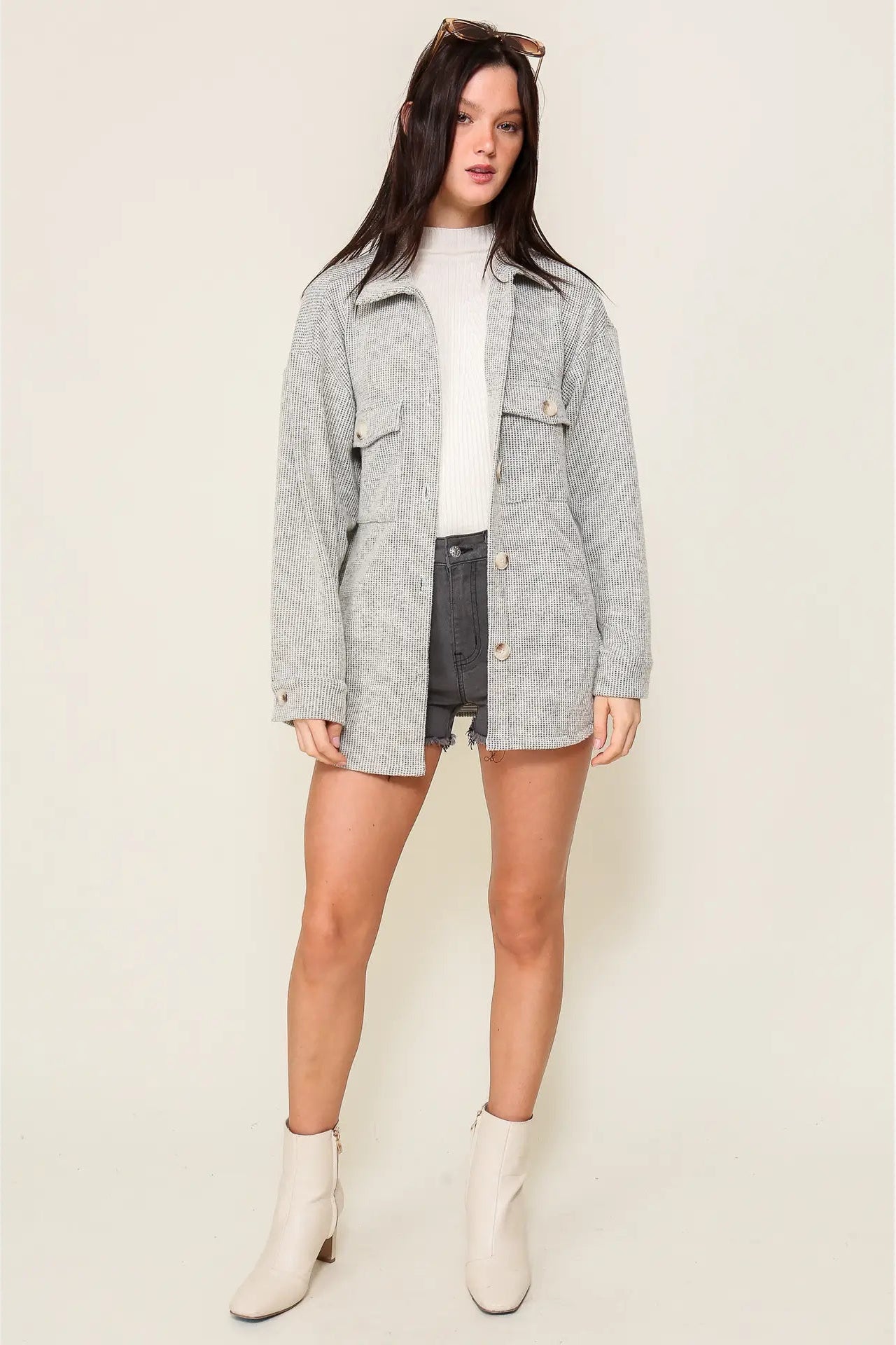 A woman modeling a light gray heathered oversized utility jacket over a white top and dark gray shorts, paired with off-white ankle boots, against a solid light beige studio background.