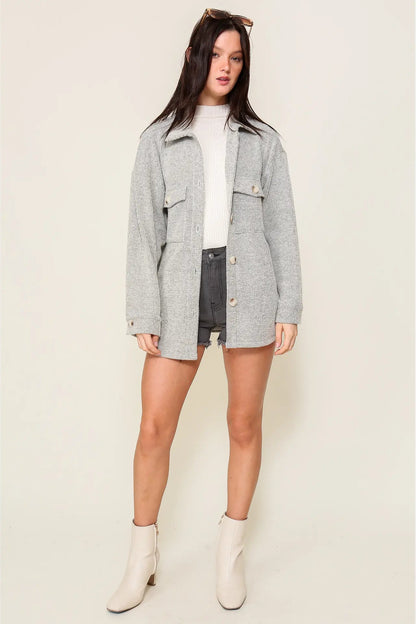 A woman modeling a light gray heathered oversized utility jacket over a white top and dark gray shorts, paired with off-white ankle boots, against a solid light beige studio background.