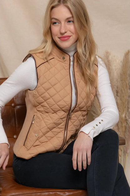 A woman wearing a tan diamond-quilted zip-front vest over a white long-sleeve top and black pants, seated on a brown leather chair against a neutral indoor backdrop.