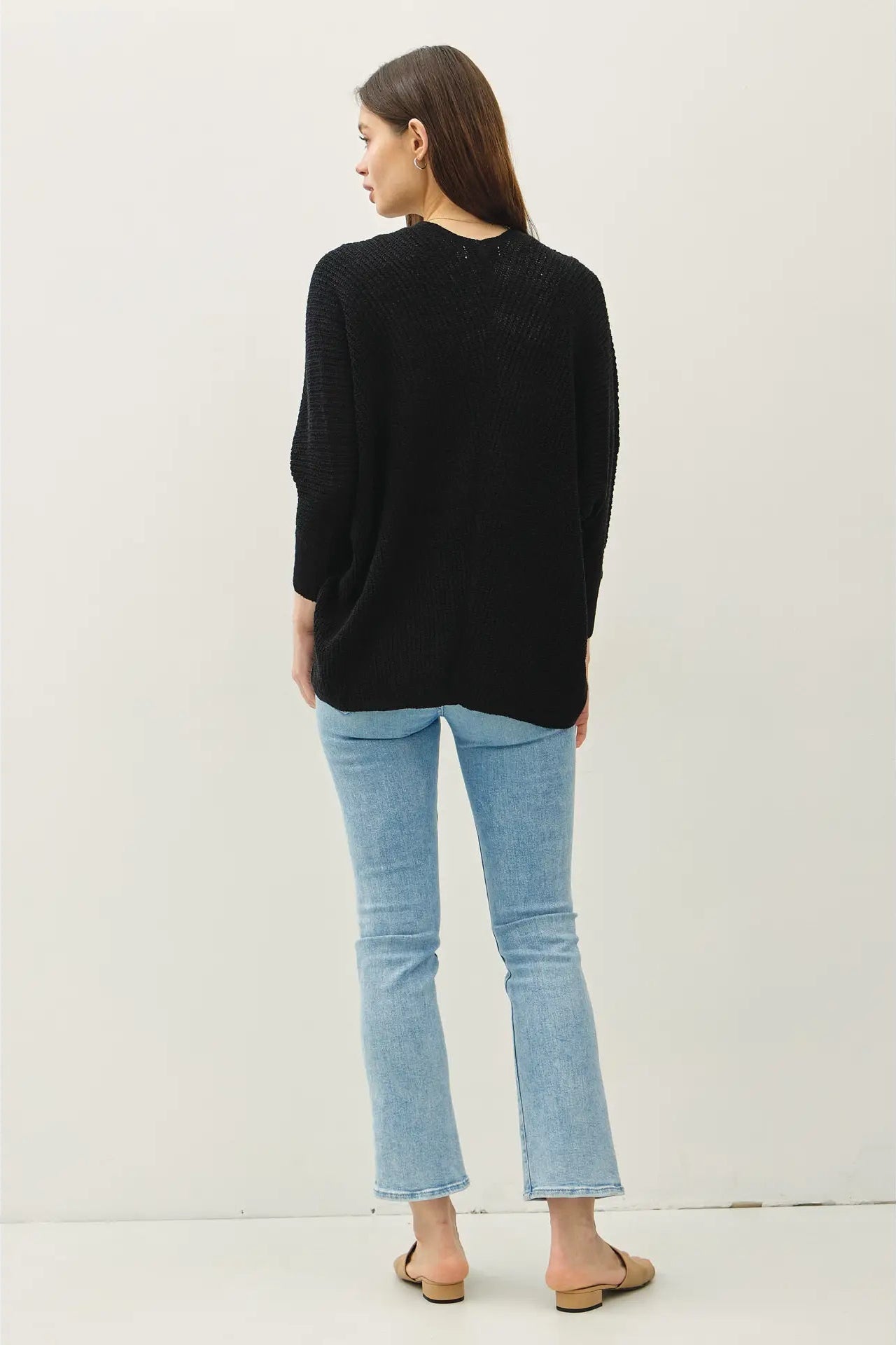 A woman is shown from the back wearing a black knit sweater, light blue cropped flare jeans, and tan flat sandals against a plain off-white studio background.