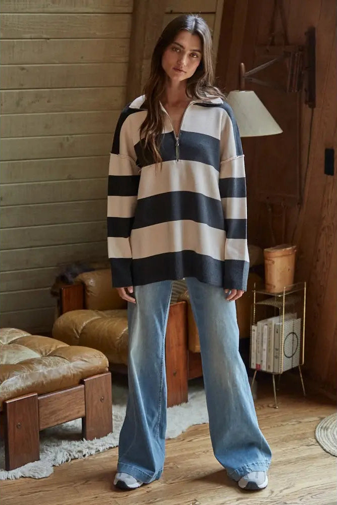 A woman stands in a rustic room wearing a long-sleeve quarter-zip pullover with bold horizontal navy and cream stripes, paired with light-wash wide-leg jeans.