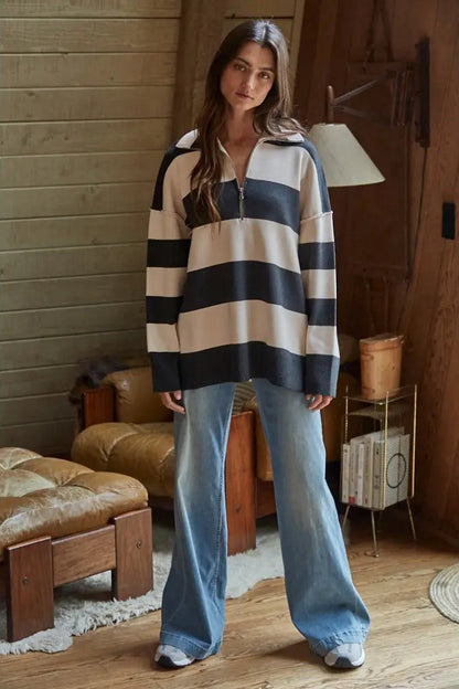 A woman stands in a rustic room wearing a long-sleeve quarter-zip pullover with bold horizontal navy and cream stripes, paired with light-wash wide-leg jeans.