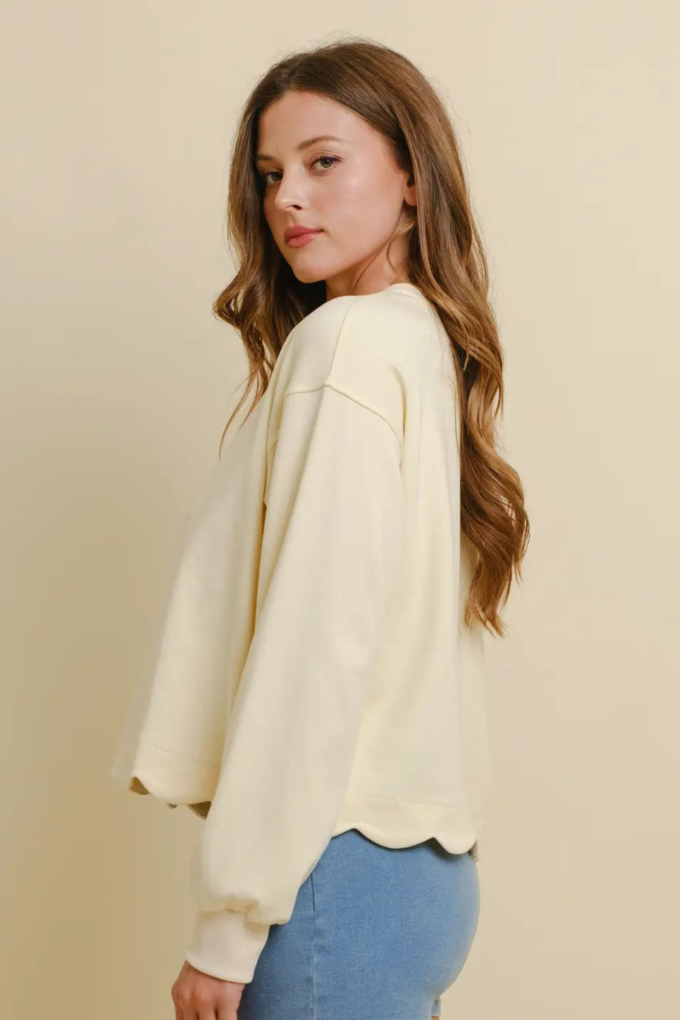 A woman with long wavy brown hair is shown from the back-three-quarter view, wearing a cream-colored oversized sweatshirt and light blue jeans against a solid beige studio background.