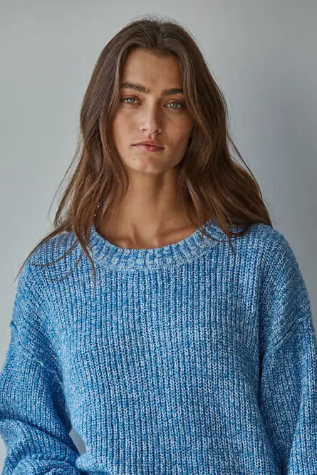A woman with long brown hair wearing a chunky, textured blue knit sweater, photographed against a neutral gray studio background.