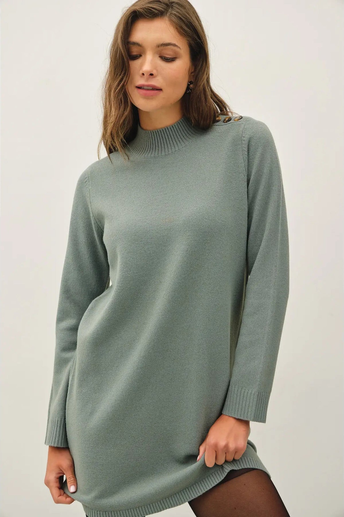 A woman wearing a sage green ribbed knit sweater dress, photographed against a neutral light gray studio background.