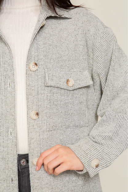 A close-up of a person wearing a light gray textured tweed button-up shirt jacket over a white ribbed top, with a dark belt visible at the bottom.