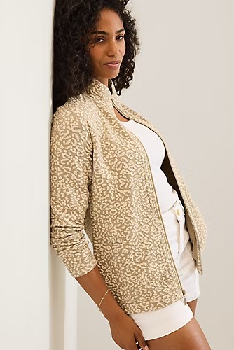 A woman wearing a beige and white patterned zip-up jacket over a white top and white shorts, leaning against a plain off-white wall.