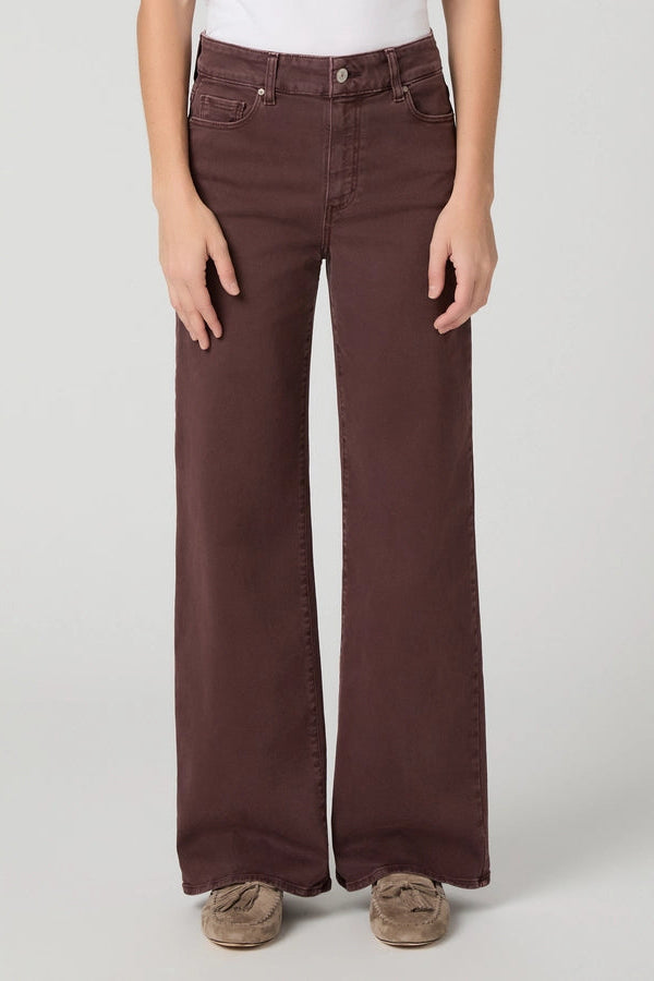 A person wearing dark brown wide-leg jeans with a white top and brown slip-on shoes, standing against a light gray studio background.