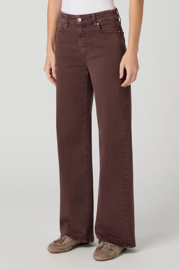 A person wearing dark brown wide-leg jeans with a white top and brown suede shoes, standing against a light gray studio background.