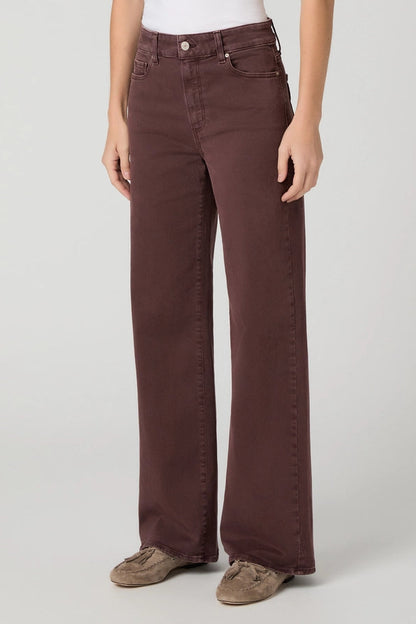 A person wearing dark brown wide-leg jeans with a white top and brown suede shoes, standing against a light gray studio background.