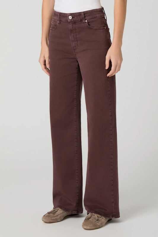 A person wearing dark brown wide-leg jeans with a white top and brown suede shoes, standing against a light gray studio background.