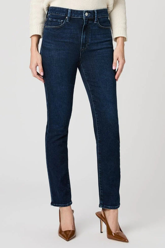 A model wearing dark blue slim-fit jeans, a cream knit sweater, and brown pointed-toe heels, shown from the waist down against a light gray studio background.