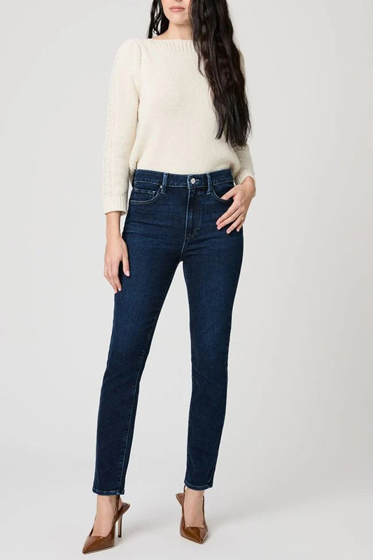 A woman modeling a pair of dark blue skinny jeans with a cream knit sweater and brown pointed-toe heels, standing against a light gray studio background.