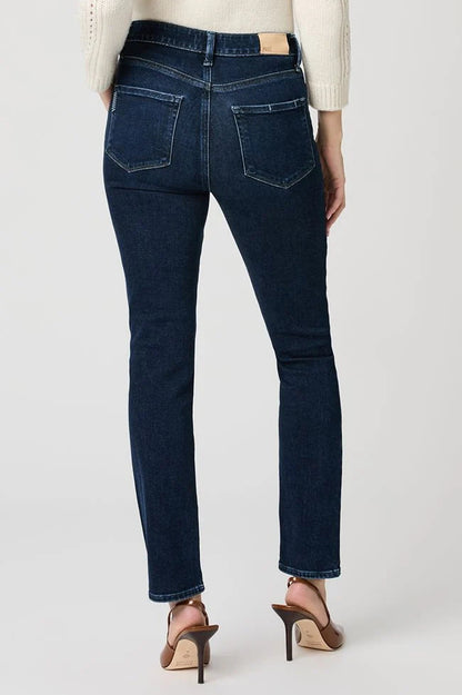 Rear view of a model wearing dark blue slim-fit jeans with a beige leather patch on the back waistband, styled with a cream sweater and brown high-heeled sandals against a light gray studio background.