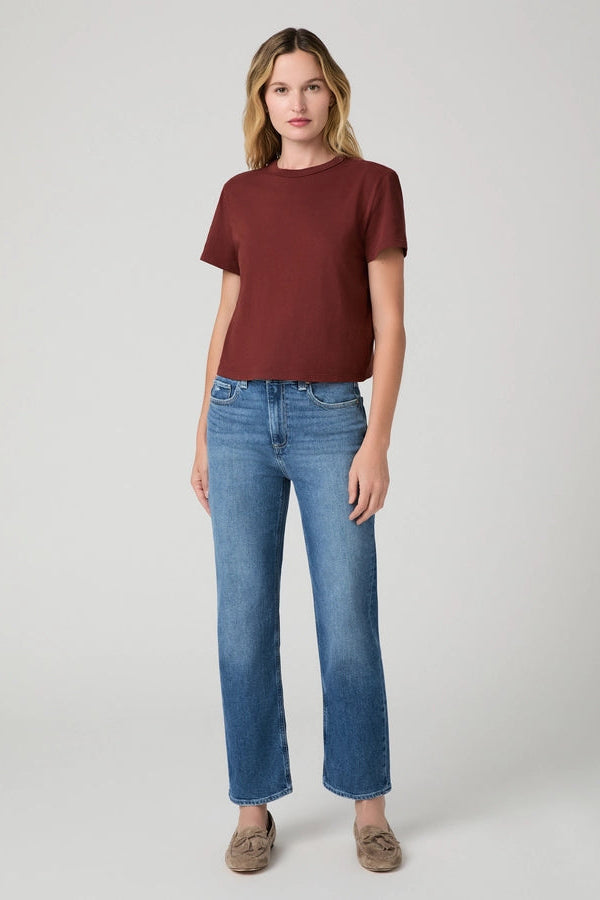 A woman modeling a short-sleeve crewneck t-shirt in burgundy and blue jeans, standing against a neutral light gray studio background.