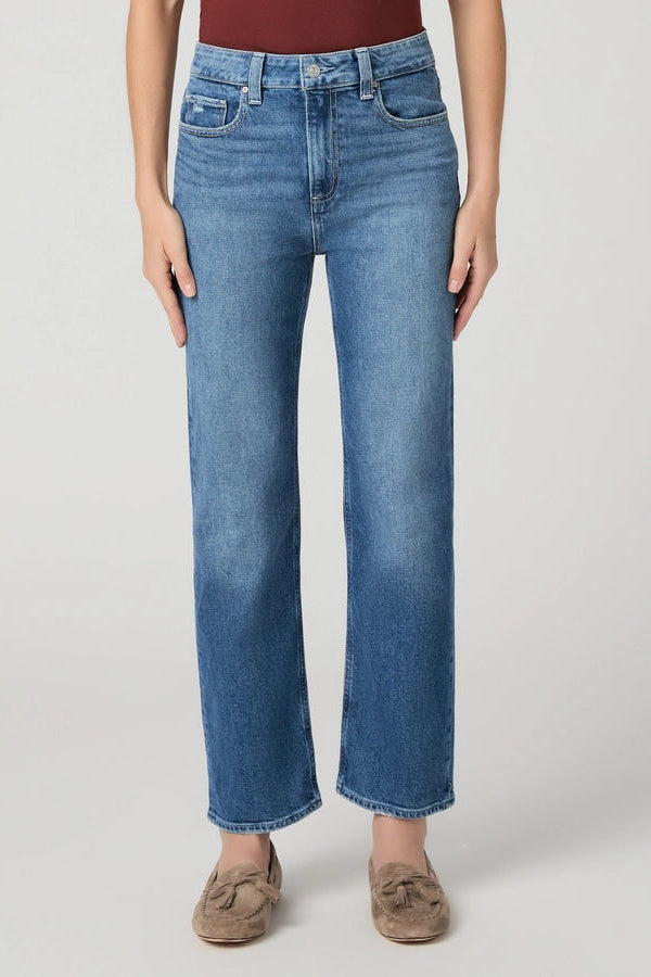 A model wearing a pair of medium-wash blue straight-leg jeans, styled with a dark red top and brown suede loafers, photographed against a neutral light gray studio background.