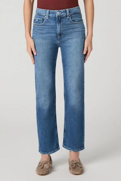 A model wearing a pair of medium-wash blue straight-leg jeans, styled with a dark red top and brown suede loafers, photographed against a neutral light gray studio background.