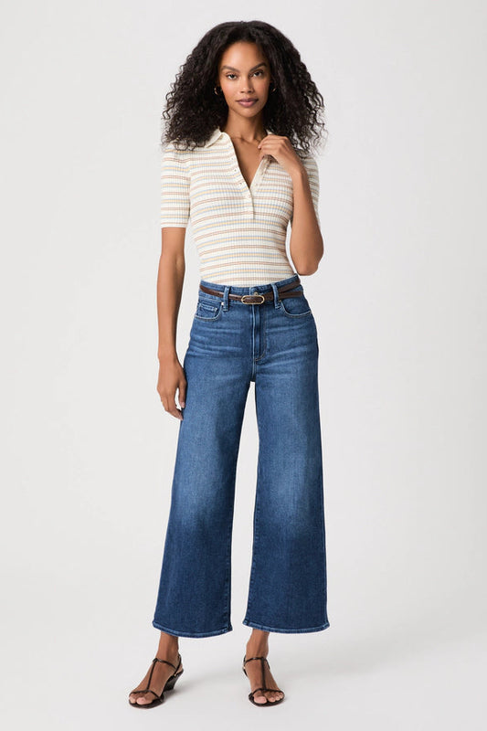 A woman modeling a cream and navy striped short-sleeve polo shirt paired with high-waisted, wide-leg blue jeans and brown strappy sandals, photographed against a solid light gray studio background.