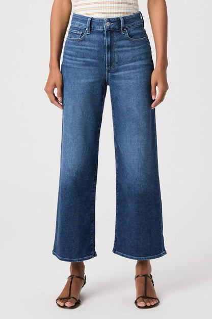 A model wearing a pair of high-waisted, wide-leg cropped blue denim jeans with a faded wash, paired with brown strappy sandals and a striped top, against a plain white studio background.