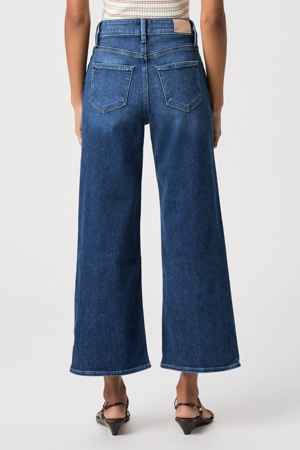 A model shown from the back wearing a pair of wide-leg cropped blue denim jeans, paired with a striped top and black sandals, against a plain light gray studio background.
