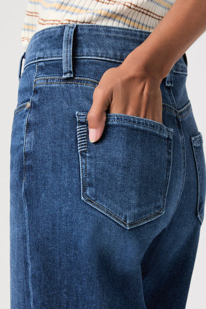 Close-up of a person wearing blue denim jeans, showing the back pocket with a hand inserted, against a light gray background.
