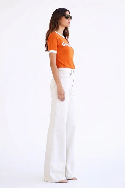 A woman modeling a bright orange short-sleeve ringer T-shirt with white trim and a white 'G' logo on the chest, paired with high-waisted white wide-leg jeans, against a plain white studio background.