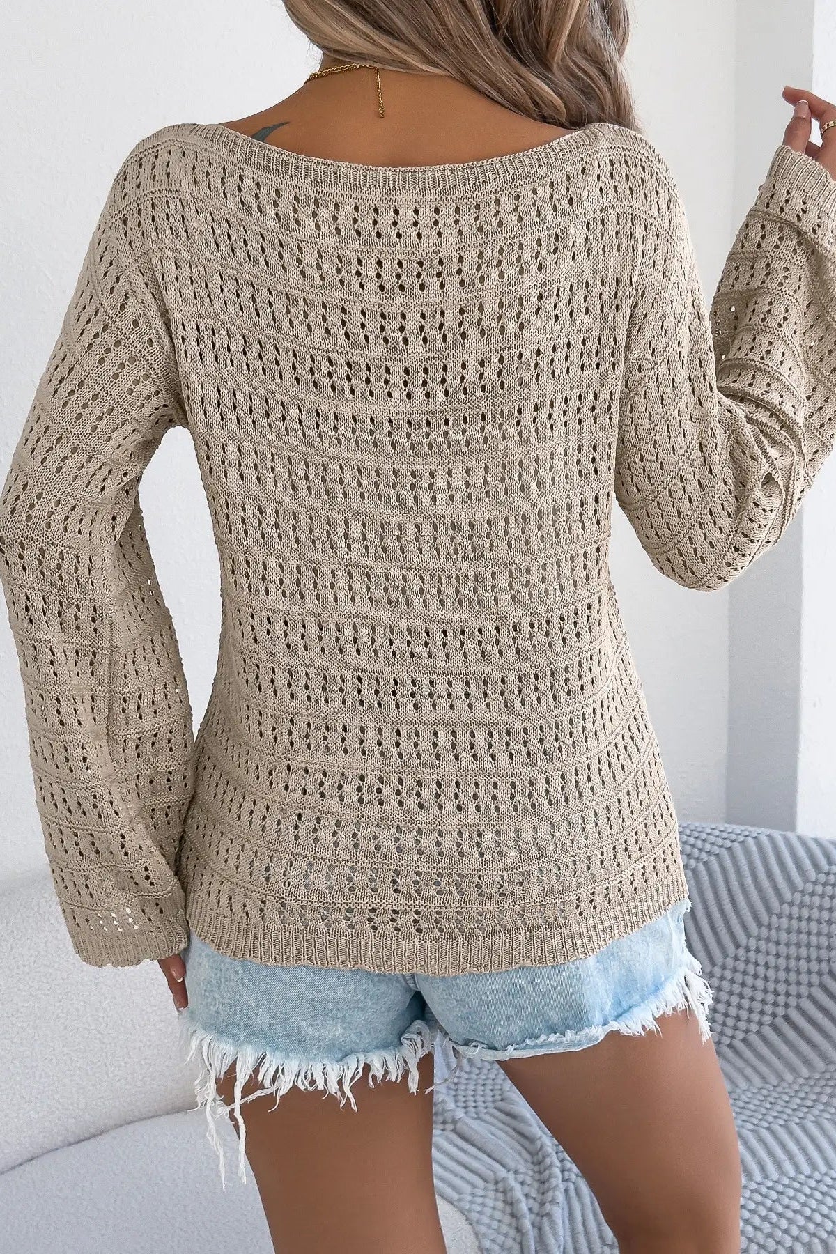 Person wearing a beige crochet sweater and denim shorts in a minimal indoor setting.