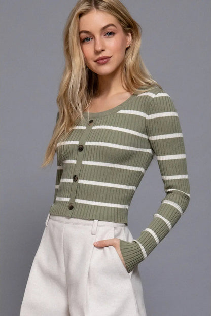 A woman modeling a ribbed knit long-sleeve cardigan with horizontal olive green and white stripes, paired with white trousers, against a solid gray studio background.