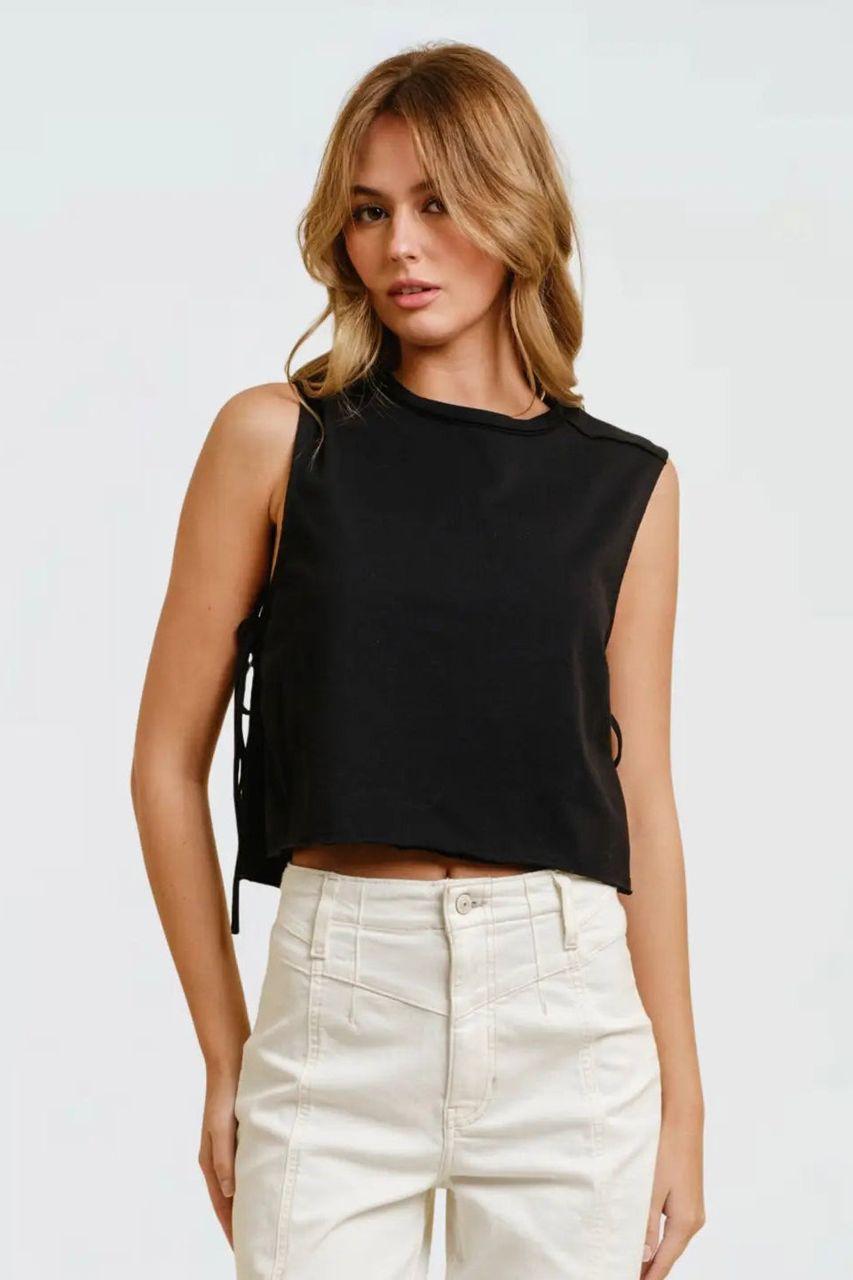 A woman modeling a sleeveless black crop top with side tie details and white high-waisted pants against a light gray studio background.