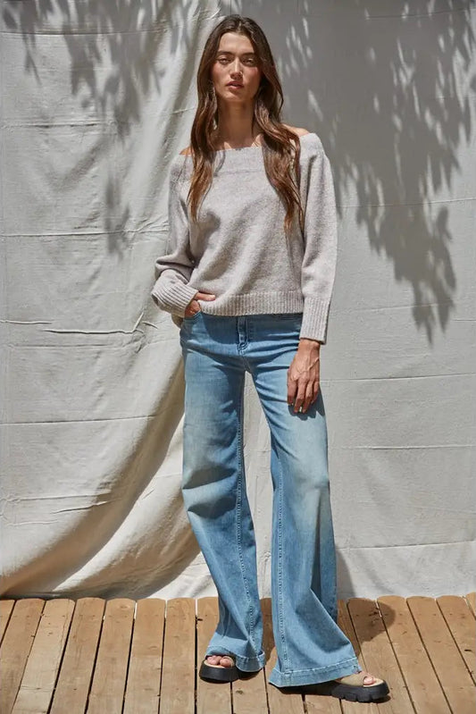 A woman stands outdoors on a wooden deck against a draped fabric backdrop, wearing a light gray knit sweater and light blue wide-leg jeans with black sandals.