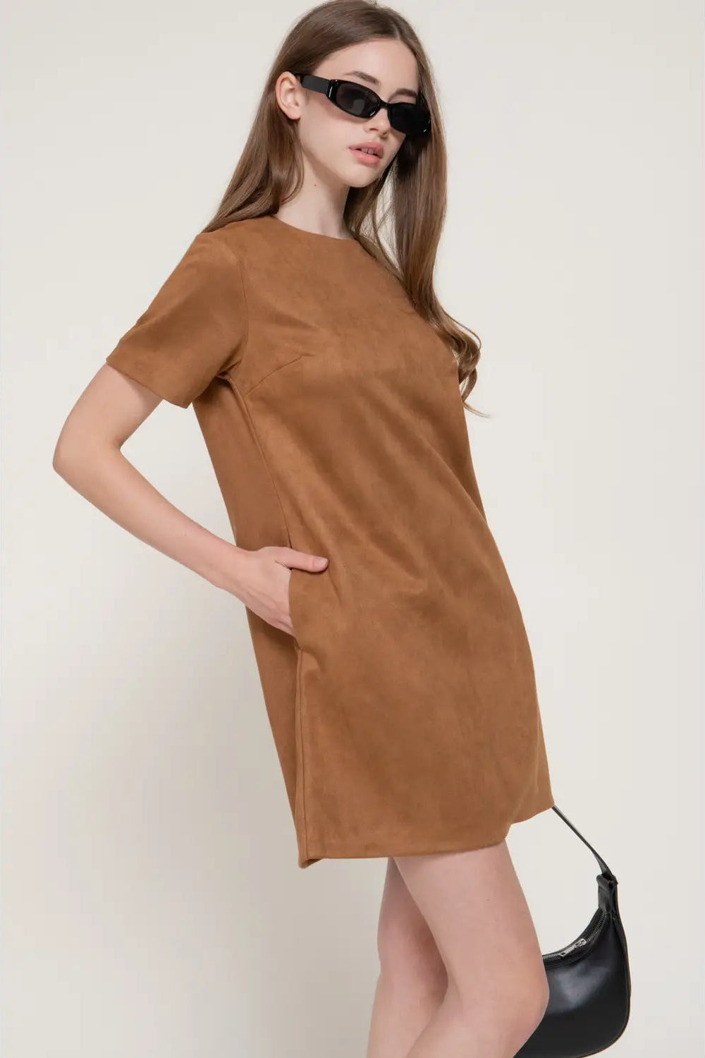 A woman modeling a short-sleeve brown dress with a relaxed fit and side pockets, styled with black sunglasses and a black crescent shoulder bag against a plain light beige background.
