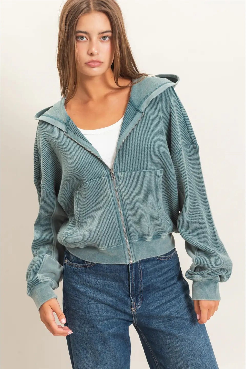 A woman modeling a sage green ribbed zip-up hoodie with textured side panels, worn over a white tank top and blue jeans, against a plain beige background.