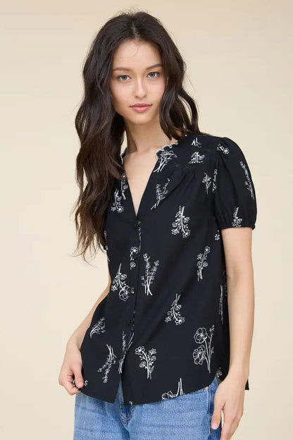 A woman wearing a black short-sleeve blouse with a white floral print, paired with blue jeans, against a solid beige background.